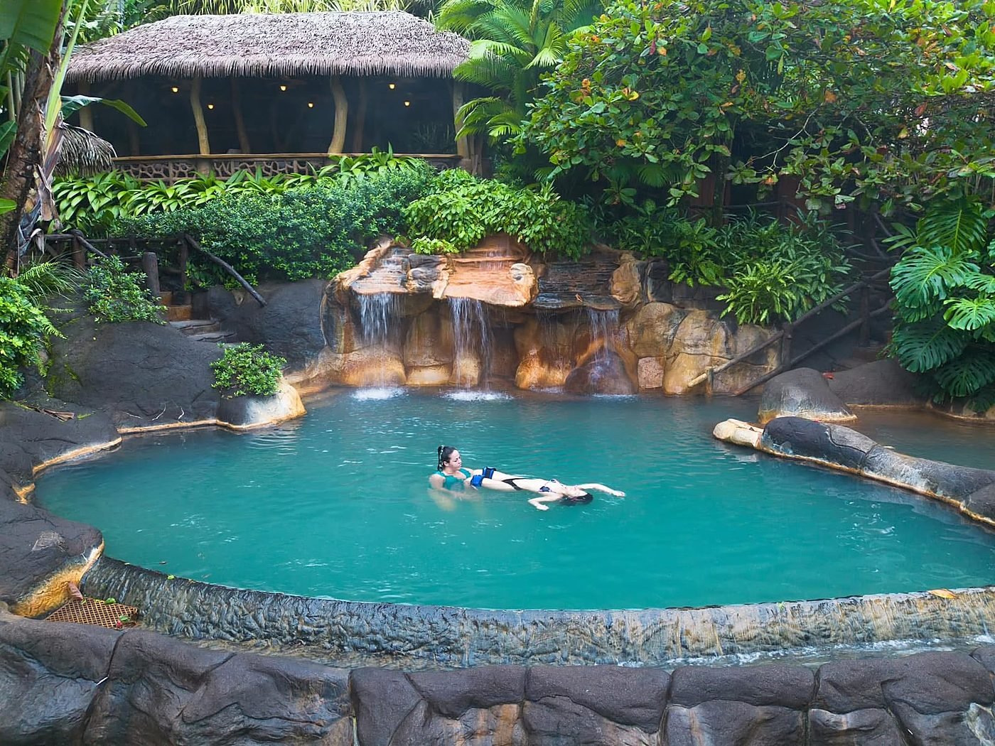Janzu session in a turquoise hot-spring pool with a waterfall and a thatched-roof lodge